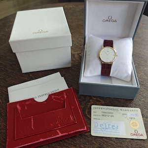Puede incluir: Un reloj Omega dorado con esfera blanca y correa de cuero burdeos, presentado en su caja original. El reloj se exhibe sobre un cojín blanco dentro de una caja gris. Incluye caja, tarjeta de garantía y folleto de instrucciones.