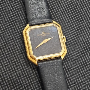 Op de afbeelding: Een Baume & Mercier Geneve horloge met een zwarte wijzerplaat en een goudkleurige bezel. Het horloge heeft een rechthoekige wijzerplaat met gouden wijzers en een zwarte leren band. Klassiek ontwerp.