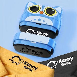 May include: A blue owl-shaped self-inking stamp featuring a white belly and large yellow eyes. The stamp mechanism displays a black dinosaur graphic with the text Kenny 123456, which is also printed on a yellow fabric collar and a black fabric collar.