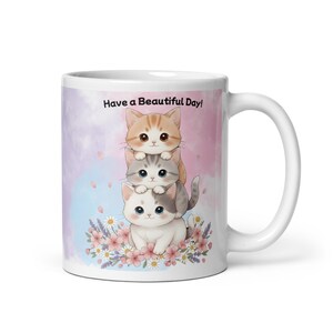 May include: White ceramic mug featuring a stack of three cartoon kittens with big eyes. The mug has a pastel background and the text "Have a Beautiful Day!" is printed on the front. Floral accents are at the bottom.