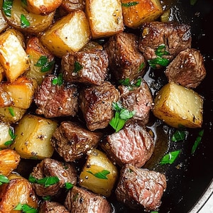 Op de afbeelding: Close-up van een pan gevuld met gekookt voedsel. Het gerecht bevat goudbruin gebakken aardappelen en aangebraden rundvleesblokjes, gegarneerd met verse groene kruiden. Het eten glanst met kookvocht, wat wijst op een hartige maaltijd.