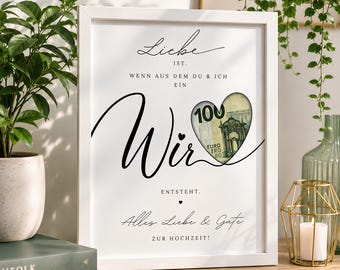 May include: A white rectangular frame containing a print with black German text that reads Liebe ist, wenn aus dem Du & Ich ein Wir entsteht, Alles Liebe & Gute zur Hochzeit. A heart-shaped cutout reveals a 100 Euro banknote behind the paper.