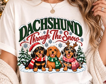 May include: A white t-shirt featuring a graphic of three dachshunds in the snow with the text "DACHSHUND Through The Snow". The dogs wear winter accessories including a Santa hat, a green scarf, and reindeer antlers with scarves.