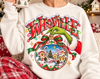 May include: A white long-sleeve sweatshirt featuring a graphic of a green hand holding a Christmas ornament with a Whoville village scene inside. The word WHOVILLE is printed in red above the ornament, surrounded by colorful holiday lights.