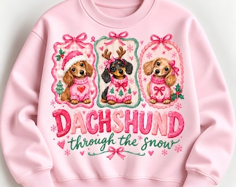May include: A light pink crewneck sweatshirt featuring three dachshunds in festive sweaters and holiday accessories within decorative frames. The text reads DACHSHUND through the snow, accented with pink bows, hearts, and snowflakes.