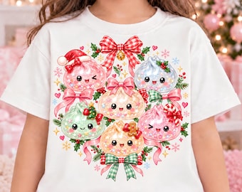 May include: A white t-shirt featuring a central graphic of five smiling, glittery dumpling characters arranged in a circle. The design includes red and green plaid bows, a Santa hat, a small Christmas tree, a gingerbread man, and candy canes.