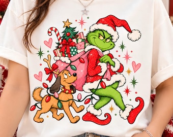 May include: A white t-shirt featuring a graphic of a green character in a red Santa suit carrying a pink sack filled with gifts and a Christmas tree. A brown dog with reindeer antlers and a string of colorful Christmas lights stands nearby.
