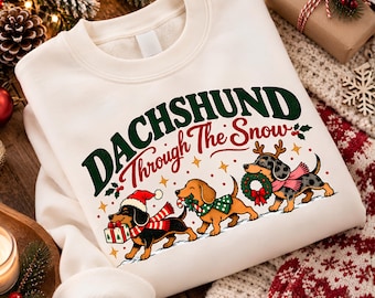 May include: A cream-colored crewneck sweatshirt featuring the text "DACHSHUND Through The Snow" in dark green and red. The graphic includes three dachshunds wearing holiday accessories, carrying a gift, a candy cane, and a wreath.