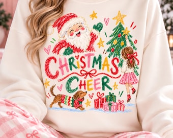 May include: A cream-colored sweatshirt featuring a colorful crayon-style graphic of Santa Claus, a decorated Christmas tree, a child placing a star, and a dachshund in a sweater. The text reads Christmas Cheer, surrounded by hearts and gifts.