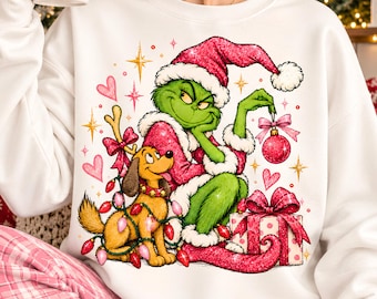 May include: A white long-sleeve sweatshirt featuring a graphic of a green character in a pink Santa hat and coat, holding a pink ornament. Beside the character is a brown dog wrapped in pink and red Christmas lights, sitting next to a gift.