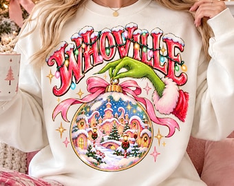 May include: A cream-colored crewneck sweatshirt featuring a graphic print of a green hand holding a Christmas ornament. The ornament contains a snowy village scene, and the text "WHOVILLE" is written in large red letters above the ornament.