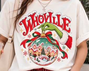 May include: A white t-shirt featuring a graphic of a green hand holding a Christmas ornament. Inside the ornament is a snowy village scene with a tree. The word WHOVILLE is printed in large red letters above the hand, accented with white snow.