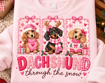 May include: A light pink sweatshirt featuring three dachshunds in individual frames. The dogs wear winter accessories like hats and scarves. Below the illustrations, the text reads "DACHSHUND through the snow" in pink and red lettering.