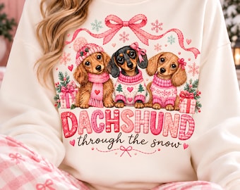 May include: A cream-colored sweatshirt featuring a graphic of three dachshunds wearing pink winter accessories. The text reads DACHSHUND through the snow. The design includes pink bows, snowflakes, hearts, and gift boxes in a festive style.