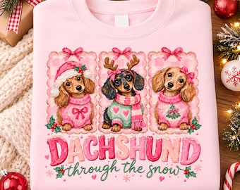 May include: A light pink sweatshirt featuring three dachshunds in rectangular frames. The dogs wear holiday sweaters, a Santa hat, and reindeer antlers. Below the dogs, the text reads "DACHSHUND through the snow" in pink and green lettering.