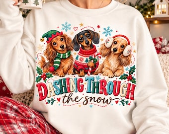 May include: A white long-sleeve sweatshirt featuring a graphic of three dachshunds in winter attire. The text reads "DASHING THROUGH the snow" in red and blue. The dogs wear a green elf hat, a red patterned sweater, and white earmuffs.