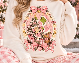 May include: A cream-colored sweatshirt featuring a central oval graphic of a green character in a pink coat and a small blonde child decorating a Christmas tree. A brown dog sits nearby, all framed by pink bows, string lights, and lace.