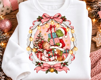 May include: A white crewneck sweatshirt featuring a central oval graphic of a child, a green creature in a Santa hat, and a puppy surrounded by Christmas gifts, a tree, and string lights, all framed by a pink ribbon and lace border.