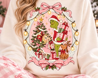 May include: A cream-colored crewneck sweatshirt featuring a central oval graphic of a green character, a young girl, and a brown dog with antlers near a decorated Christmas tree. The scene is framed by a string of lights and pink bows.