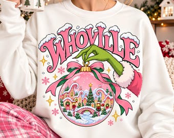 May include: A white long-sleeve sweatshirt featuring a graphic of a green hand holding a pink Christmas ornament. Inside the ornament is a snowy village scene with houses and trees. The text "WHOVILLE" is printed in large pink letters above.