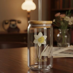 以下が含まれることがあります： 竹製の蓋とガラスストローが付いた透明なガラスタンブラー。タンブラーには、金色のスクリプトで「March」という言葉が入った水仙のデザインが施されています。タンブラーは木製の表面にあります。