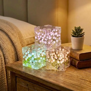May include: Three illuminated, clear cube-shaped decorative lights with embedded floral designs. The lights are arranged on a wooden nightstand, with a small potted succulent and books nearby. The cubes glow with soft colours.