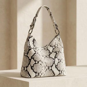 Puede incluir: Un bolso de hombro estilo hobo con un patrón de piel de serpiente en blanco y negro. El bolso tiene una sola correa sujeta con anillos de metal oscuro a ambos lados, una abertura superior curva y una base estructurada con textura de escamas.