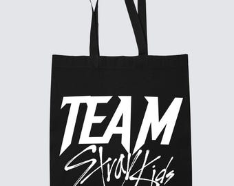 May include: Black tote bag with white text that reads "TEAM Stray Kids". The bag has two black handles and is made of a sturdy fabric. The design is a bold, graphic statement.
