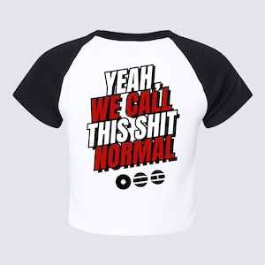 Puede incluir: Top corto blanco con mangas raglán negras. La parte trasera de la camiseta presenta el texto "YEAH WE CALL THIS SHIT NORMAL" en letras de bloque rojas, blancas y negras. Debajo del texto hay tres líneas horizontales y círculos.