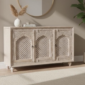 May include: A light-coloured wooden cabinet with detailed carvings and three doors. A round gold mirror hangs above the cabinet, and a potted plant stands to the right. The cabinet has a textured, distressed finish.