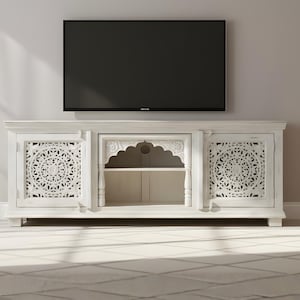 Puede incluir: Mueble de TV de madera blanca con intrincados diseños de mandala tallados en las puertas. El mueble tiene un estante abierto en el centro y está coronado por un gran televisor de pantalla plana negro. El mueble está colocado contra una pared de color neutro.