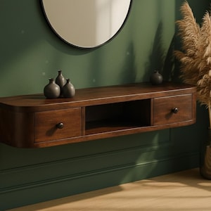 Puede incluir: Una consola flotante de madera marrón oscuro con dos cajones y un estante abierto. La consola está montada en una pared verde oscuro, con un espejo redondo encima. Jarrones decorativos y plantas secas completan la escena.