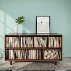 May include: A dark brown wooden record cabinet filled with vinyl records. A framed abstract art print and a vase with a large green leaf are on top. The cabinet is against a light blue wall. The cabinet is approximately 150 cm wide.