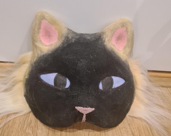 May include: A Siamese cat mask with a black face, light blue eyes, and pink nose and ears. The mask has a fluffy, cream-colored fur around the face. The mask is made of felt and fabric.