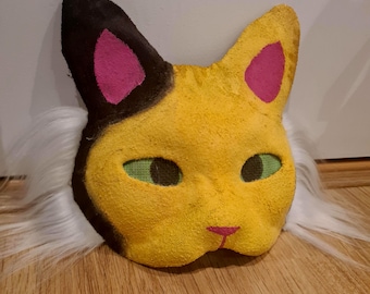 May include: A handmade cat mask with a yellow face, black ears with pink inner ears, and green eyes. White faux fur surrounds the mask. The mask is resting on a wooden surface.