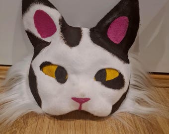 May include: A white cat mask with black markings, yellow eyes, and pink accents on the ears and nose. The mask has a fluffy white fur trim and is set against a wooden surface.