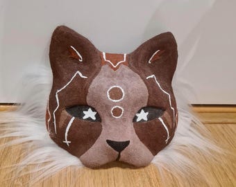 May include: A brown cat mask with white accents and star-shaped eyes. The mask features a symmetrical design with a central circle pattern and is set against a fluffy, white background.