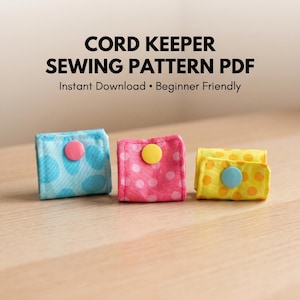 May include: Three handmade cord keepers in blue, pink, and yellow, each with a polka dot pattern and a button closure. The text "CORD KEEPER SEWING PATTERN PDF" is displayed above the cord keepers.