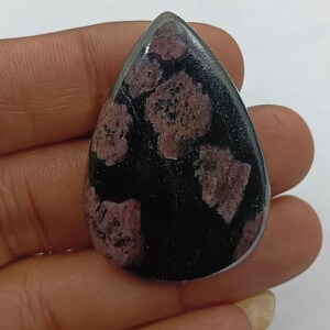 May include: A polished, teardrop-shaped gemstone. The stone has a black base with irregular patches of deep pink and purple. The gemstone is held in a hand, showing its size and shape. The stone is likely for jewellery.