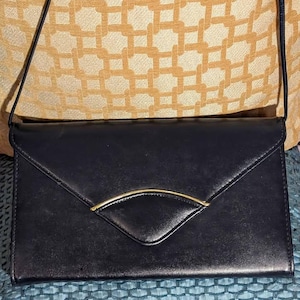 May include: A black clutch purse with a gold-colored accent. The rectangular purse has a flap closure and a thin shoulder strap. The gold accent is a curved, half-moon shape. The purse is made of a smooth material.