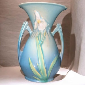 May include: A light blue ceramic vase with a flared top and two handles. The vase features a white and yellow floral design with green leaves. The vase is approximately 10 inches tall.