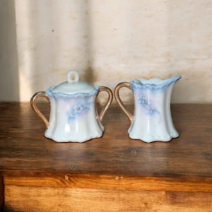 May include: A vintage porcelain sugar bowl with a lid and a creamer. Both pieces are white with a light blue scalloped edge and gold handles. Delicate blue floral accents adorn the sides of each piece. The set is displayed on a wooden surface.