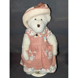 May include: A white teddy bear wearing a pink dress with floral accents and a matching hat. The dress has a Peter Pan collar and pockets with floral fabric. The hat is straw-colored with a floral embellishment.