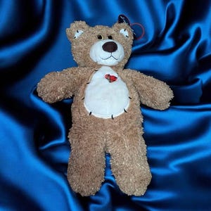 May include: A plush teddy bear with brown fur, a white belly, and a dark brown nose. The bear has a red heart-shaped patch on its chest and is set against a blue background.