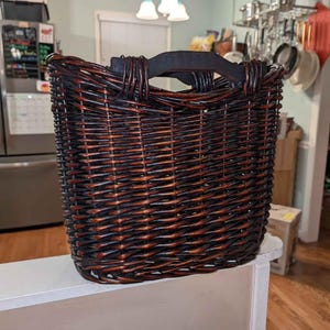 May include: A rectangular woven basket with a dark brown and reddish-brown color scheme. The basket has a black handle and is sitting on a white surface. The basket is likely used for storage or decoration.