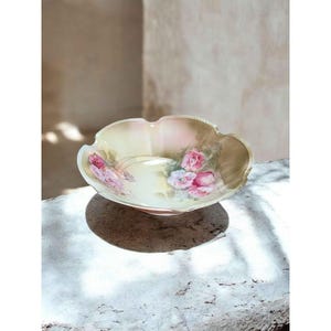 May include: A decorative, scalloped-edge bowl with a pale yellow interior and a pink and peach exterior. The bowl features hand-painted pink roses and green leaves. The bowl is sitting on a stone surface.