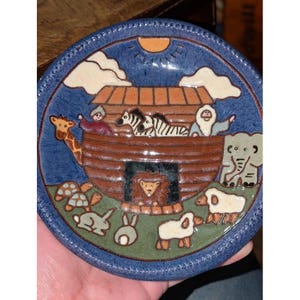 May include: A ceramic plate with a blue rim and a scene of Noah's Ark. The ark is brown, with animals and people inside. The background is blue with a sun and clouds. The plate has a diameter of approximately 6 inches.