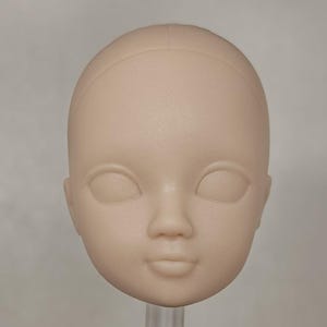 Peut inclure: Une tête de poupée vierge en matériau beige clair. La tête présente des orbites fermées, un petit nez et une bouche légèrement souriante. La tête est lisse et semble être faite d'un matériau solide. Il s'agit d'une fourniture artisanale.