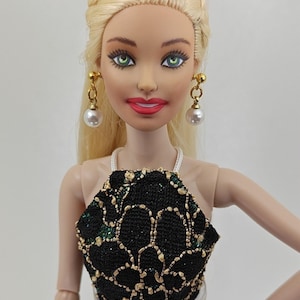 Peut inclure: Poupée blonde coiffée en tresse, vêtue d'un haut noir et doré à motifs et de boucles d'oreilles en perles. La poupée a les yeux verts et du rouge à lèvres rouge. Un bracelet doré est visible.