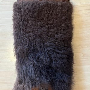 May include: A brown fur boot cover with a leather strap and buckle at the top. The boot cover has a zipper at the bottom. The fur is dark brown and covers the majority of the boot cover.
